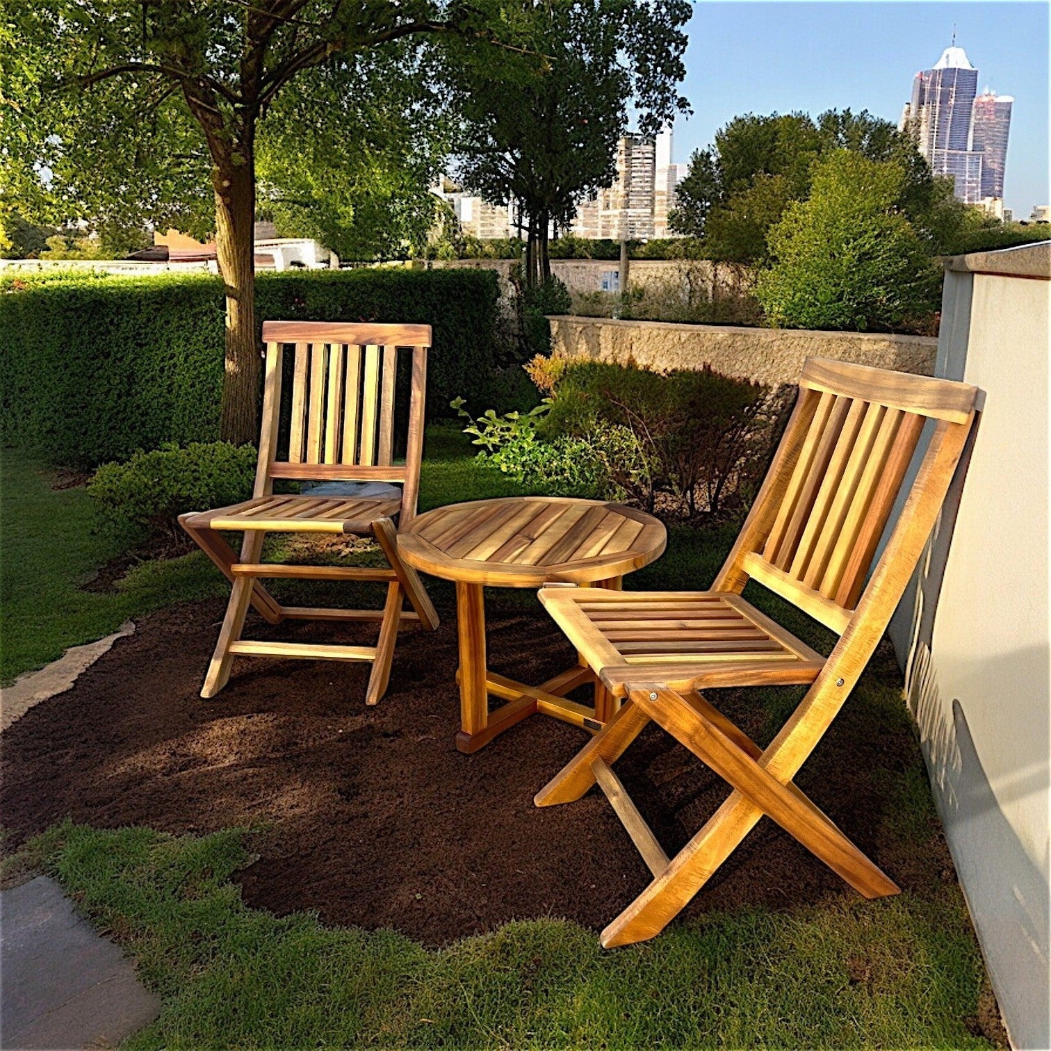 Hawaiian Discovery Furniture round acacia wood side table set outdoors between two folding wooden chairs on grass.